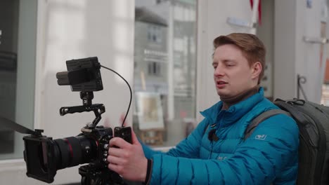 Medium-shot-of-camera-man-in-blue-jacket-directing-talent-while-holding-and-controlling-the-camera-on-a-small-town-main-street