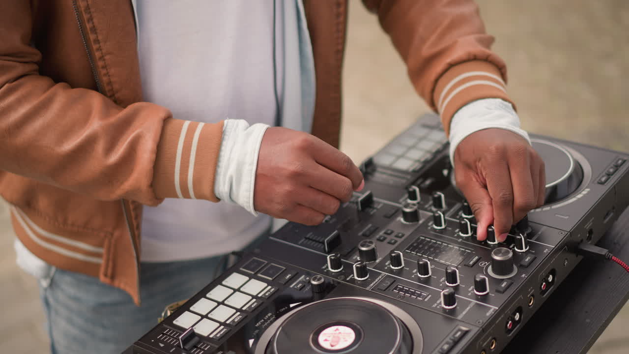 Ampie mani di un DJ nero che mixano su una console controller, musicista con giacca marrone che manipola pad e fader, esibizione di strada urbana soleggiata con un'atmosfera casual diurna, groove energico e azione di remix dal vivo