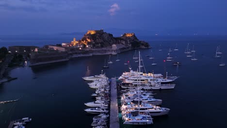 Hermosa-Vista-Aérea-Nocturna-De-La-Zona-De-Yates-De-Corfú-Y-La-Antigua-Fortaleza-Con-Templo-Griego-Iluminado-Y-Faro