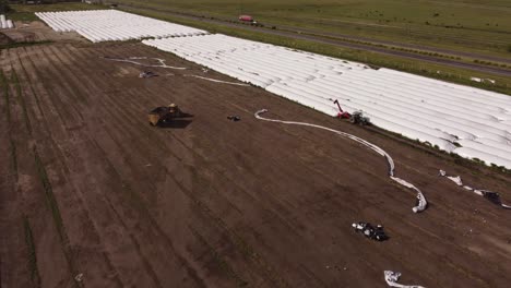 aerial flyover farm field with tractor and silo bags during harvest loading process in rural area during sunlight