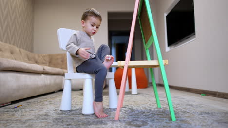 small caucasian baby sitting on the chair drops an earpiece. funny kid lifts an earpiece with toes and puts it into ear. low angle view.