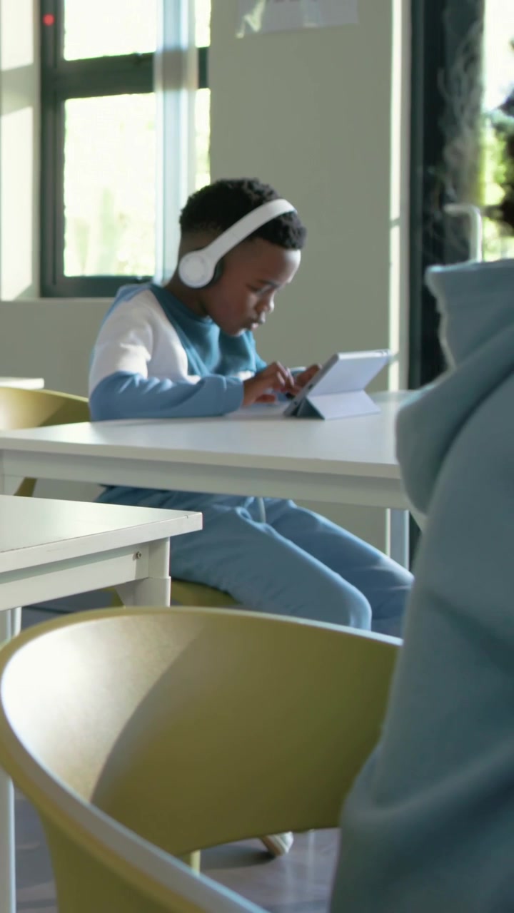 Vertical video: Tapping male student wearing headphones swiping tablet at school desk studying