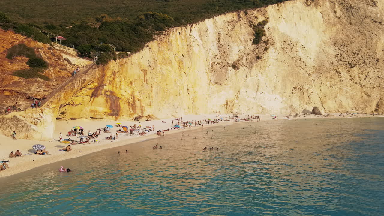 ZAKYNTHOS, GREECE - AUGUST 23, 2021: Aerial drone view of the Ionian sea coast. Rocky cliff, beach with multiple resting and swimming people, blue water. Sunset
