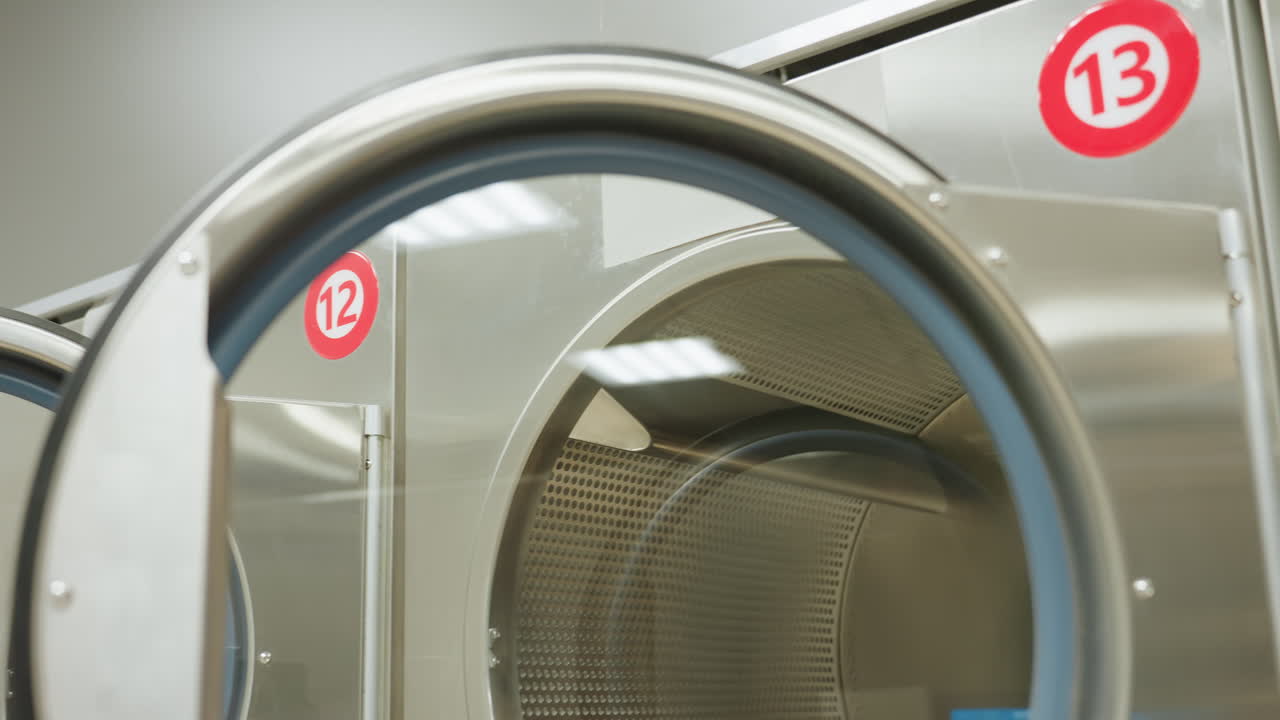 Round view of labeled industrial washer in bright laundry interior, door open revealing stainless drum, number thirteen sticker visible, modern equipment detail, reflections on metal surface