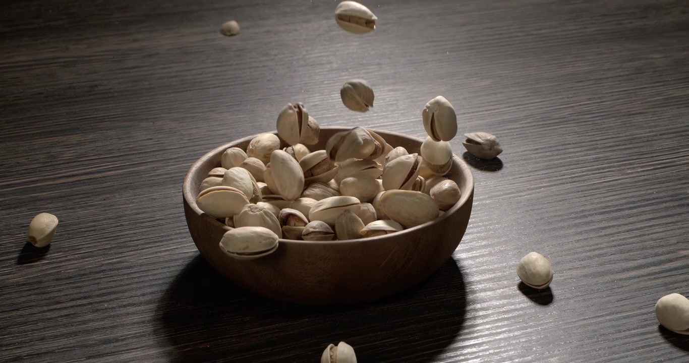 Ciotola di legno piena di pistacchi su un tavolo di legno scuro, gustosi pistacchi salati sparsi intorno, concetto di spuntino sano e cibo naturale. Ripreso con telecamera super slow motion a 1600 fps.