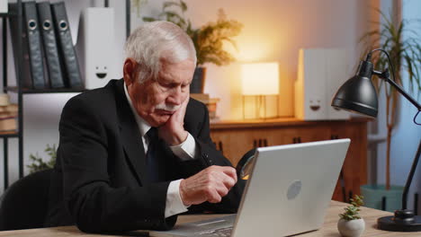 bored senior businessman working at home office holding magnifying glass looking at laptop screen