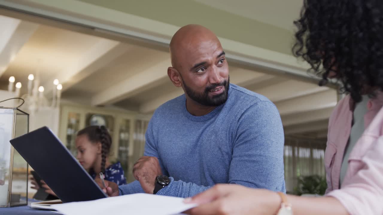 Happy biracial couple doing paperwork and using laptop at table, slow motion