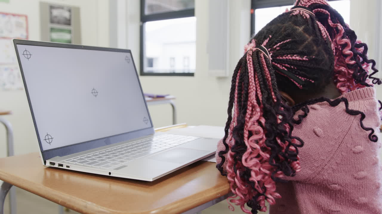 Girl with colorful braids resting head on desk beside laptop in classroom, at school, copy space