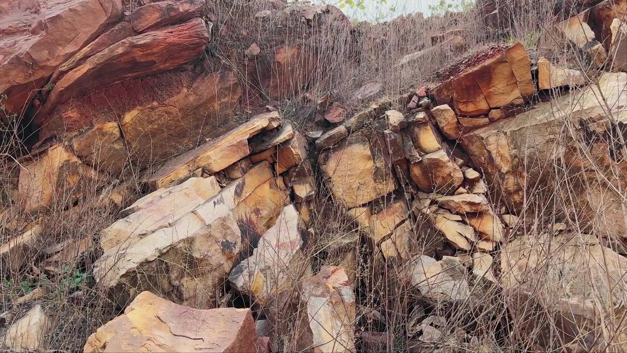 Very slow panning close up shot of fractured layered rock formations displaying weathered surfaces mineral staining and natural geological textures Interwoven dry grasses add detail to the rugged land
