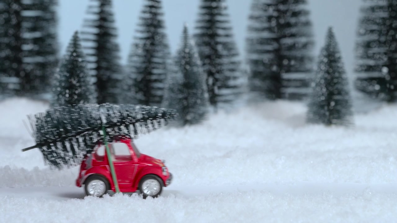 Un coche de juguete rojo con un árbol de Navidad en el techo conduce por un bosque nevado. De izquierda a derecha. Decoración navideña