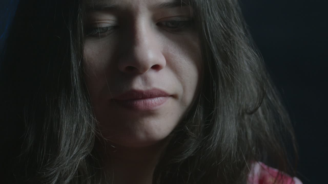 Close-up de uma jovem mulher hispânica sorrindo de lado enquanto olha para o telefone. A iluminação suave cria uma atmosfera misteriosa e íntima, realçando suas características faciais com sombras delicadas
