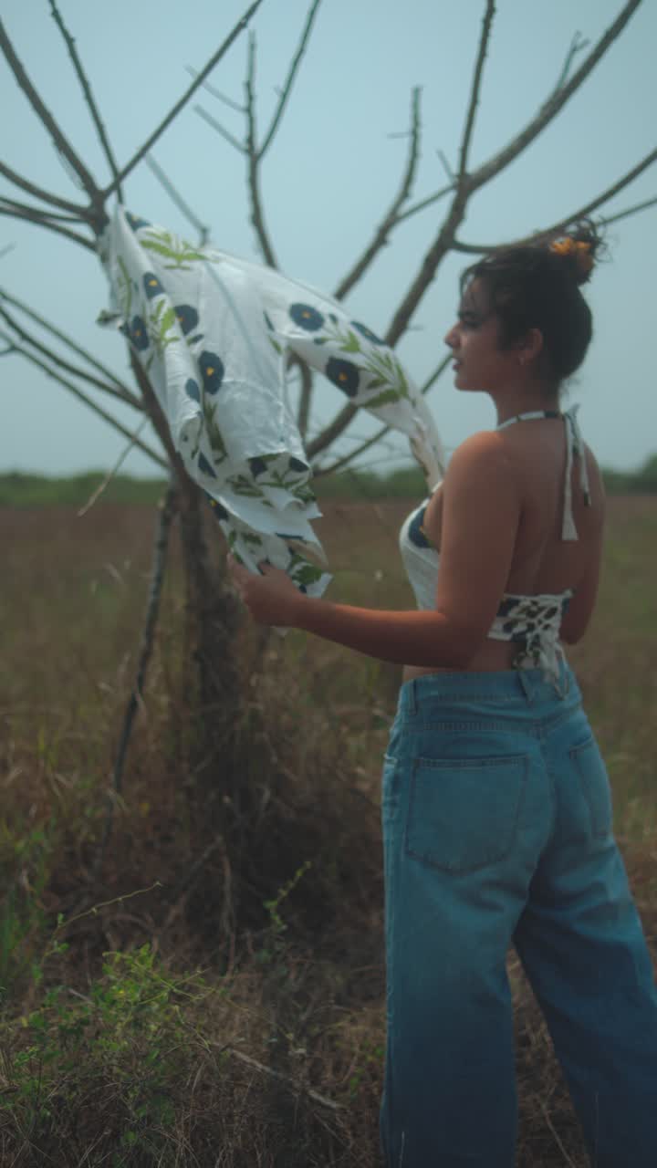 Young woman standing by a tree, playfully hanging a patterned shirt in the field