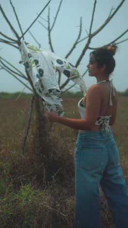 young woman standing by a tree, playfully hanging a patterned shirt in the field