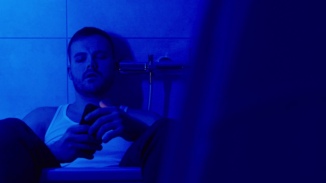 Sad young man sitting in a bathtub in a blue-lit bathroom at night, quietly using his smartphone. Concept for loneliness, mental health, insomnia, and digital overuse