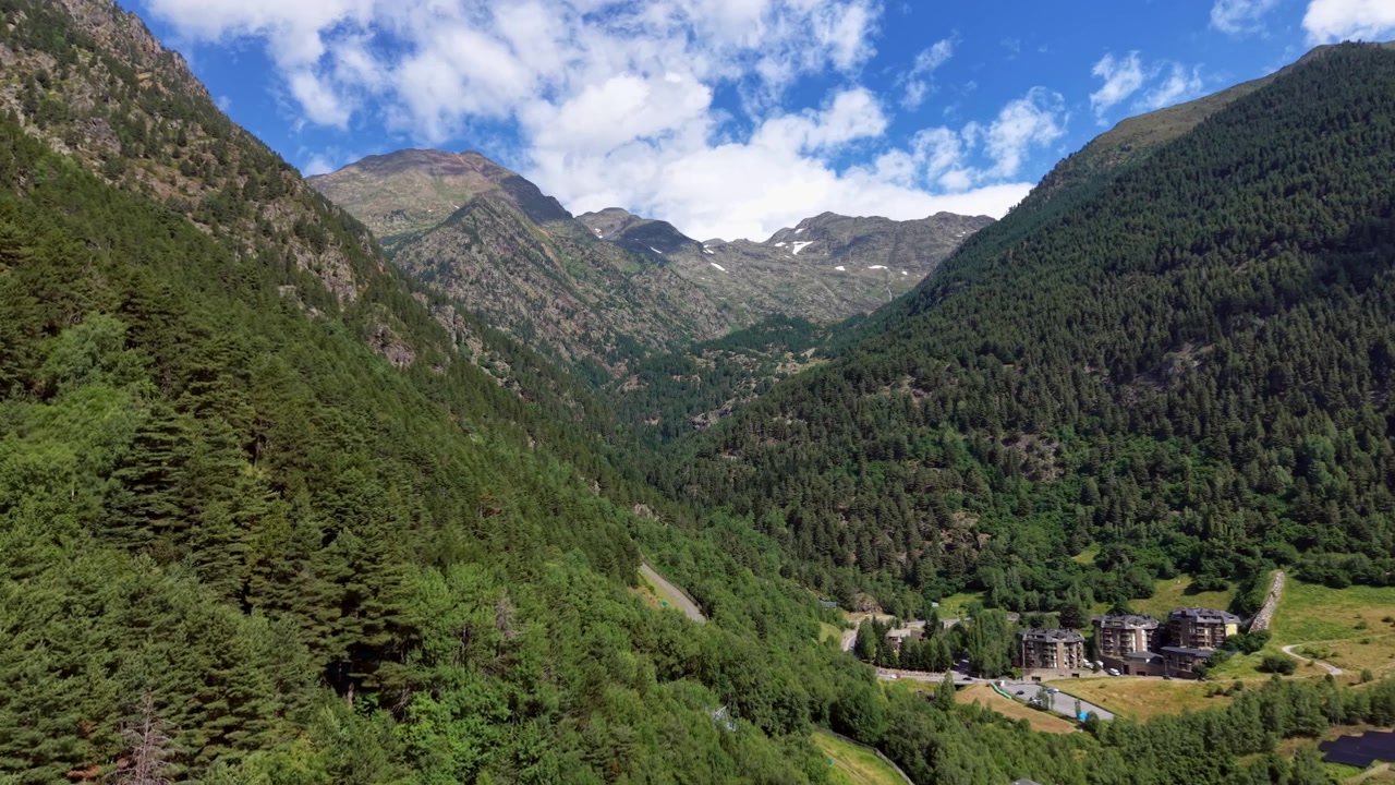 Vallée d'Arinsal en Andorre, montrant des montagnes boisées d'un vert luxuriant, des sommets enneigés des Pyrénées, une route sinueuse et des bâtiments de station nichés dans un paysage alpin sous un ciel bleu