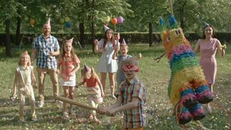 Niños-Con-Sus-Padres-En-Una-Fiesta-De-Cumpleaños-En-El-Parque,-En-Primer-Plano-Un-Niño-Rompe-Una-Piñata