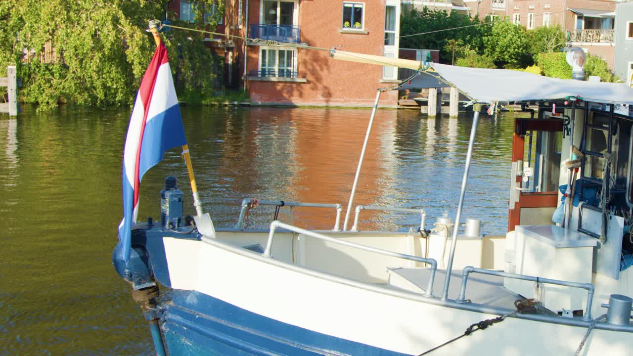 A small boat with a Dutch flag moves gently on a sunlit canal in Haarlem, with historic brick buildings and greenery in the background