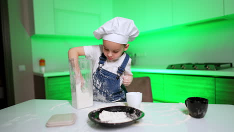 toddler boy tries to get some flour form container but spills a lot. baby chef puts some flour into the plate.