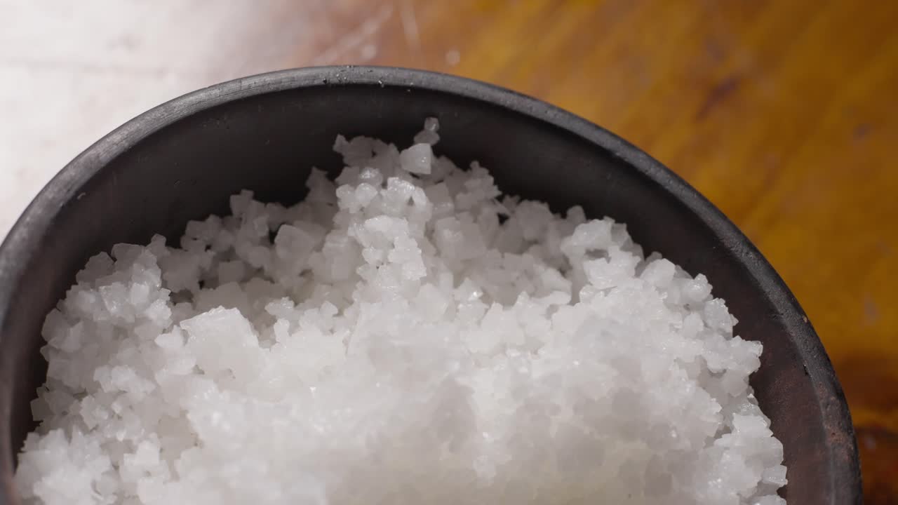 A Caucasian hand carefully takes salt crystals from a small dark salt pot in close-up view