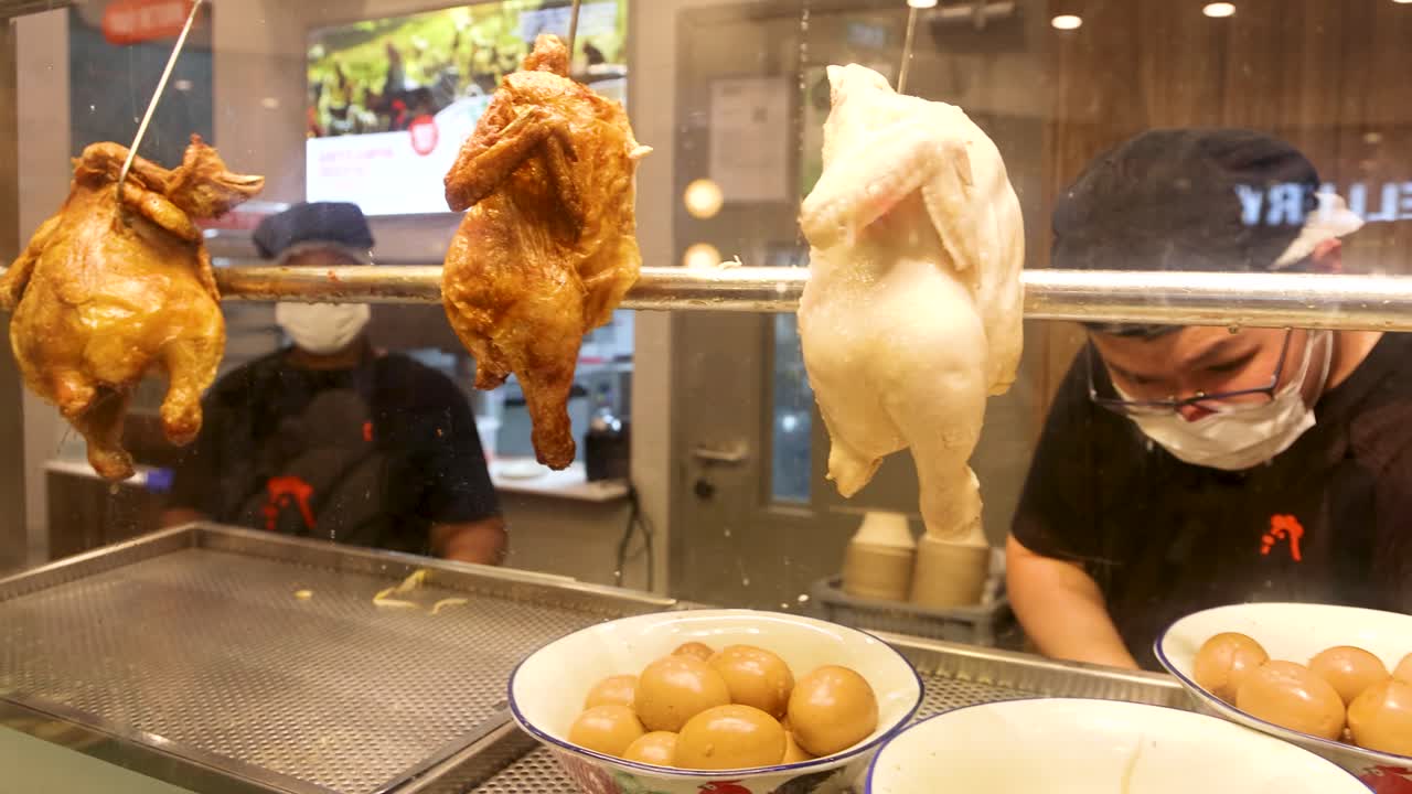 Cook in face mask works behind glass display with roasted and steamed whole chickens, eggs, and bright indoor lighting at a Singaporean food stall