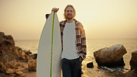 Portrait-of-a-tall-blond-guy-with-a-beard-in-a-plaid-shirt-and-a-white-T-shirt-who-stands-and-holds-his-white-surfboard-near-him-on-a-rocky-shore-near-the-sea-early-in-the-morning-at-Sunrise