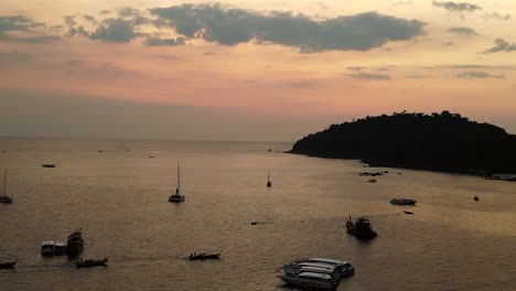 The-sun-sets-over-the-calm-ocean,-casting-a-warm-glow-on-boats-and-an-island-in-the-distance,-painting-the-sky-in-shades-of-orange-and-pink