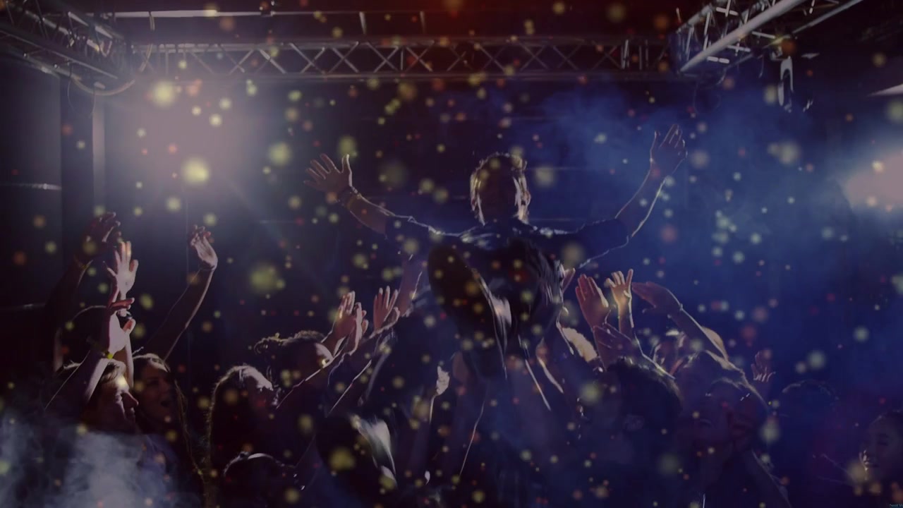 Un artiste se penche dans la foule, se faisant soulever et passer sous les lumières du concert, tandis que des confettis flottent.