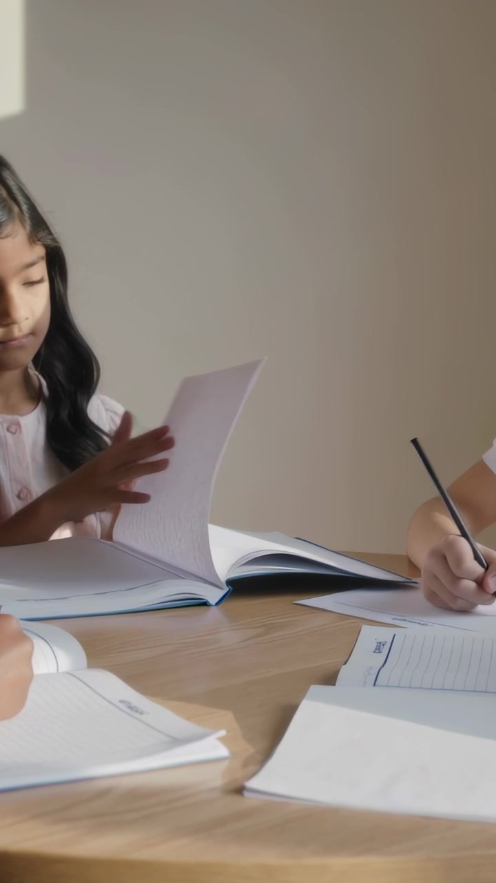 Video verticale: Ragazza indiana in camicetta rosa che inizia i compiti, sfogliando le pagine del quaderno al tavolo di casa.