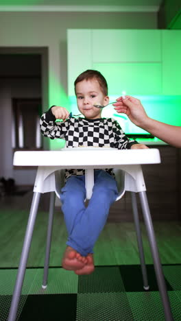 baby boy sitting at high chair eating using a fork. mother shoves the spoon to her child but he refuses. vertical video.