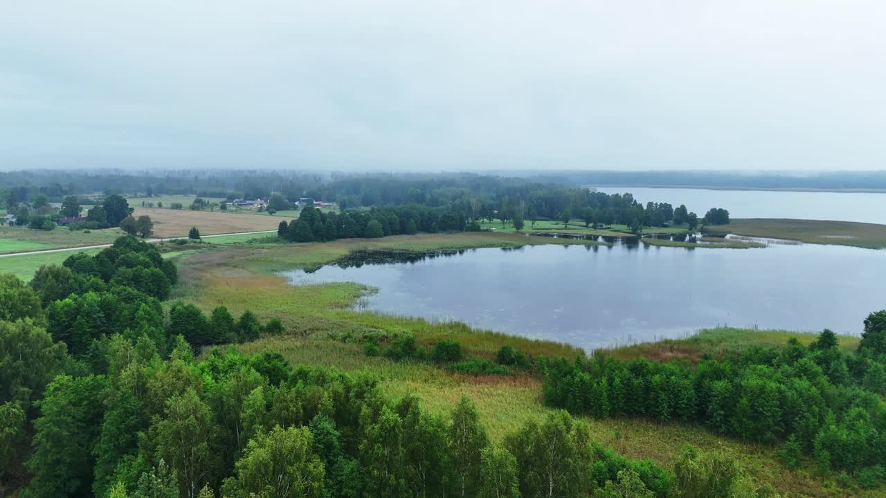 Aerial forward tracking shot moving over forest and fields towards foggy lake with misty horizon