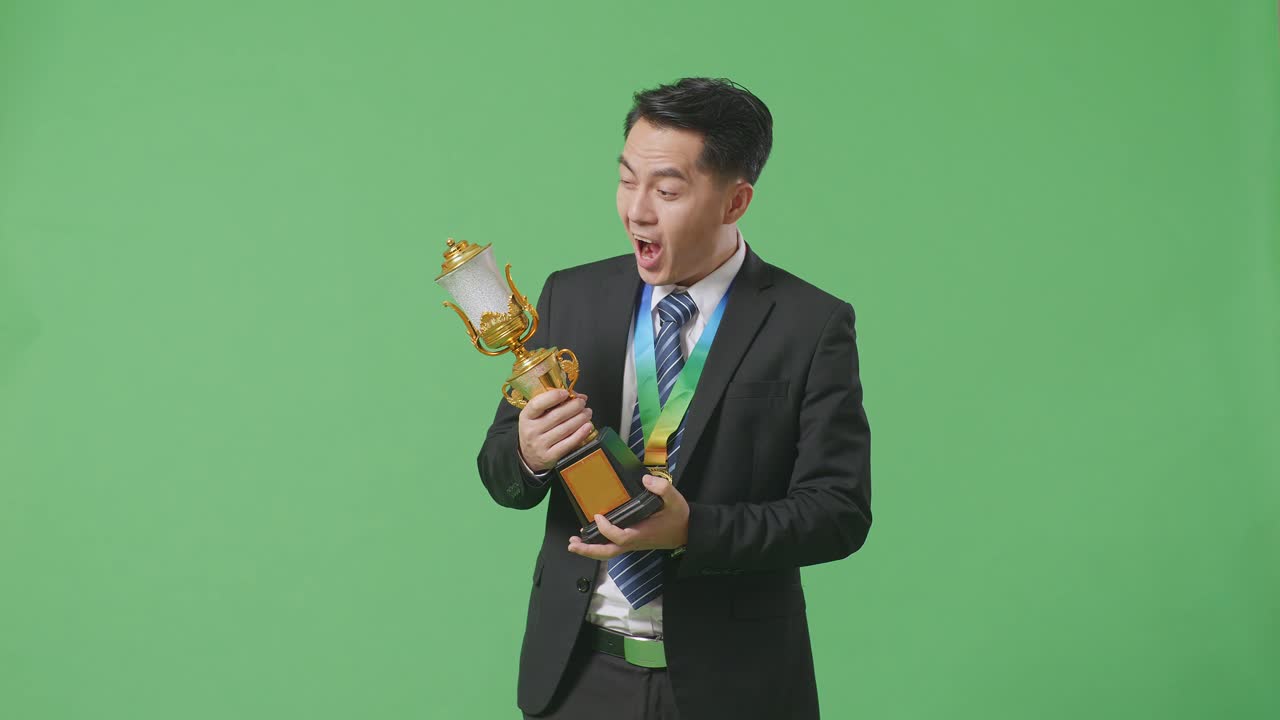 슈트와 타이를 입고 금메달을  아시아 사업가 한 명이 손에 금 트로피를 들고 와우라고 말하며 스튜디오의 녹색 스크린 배경에서 첫 번째 우승자로서 행복합니다.