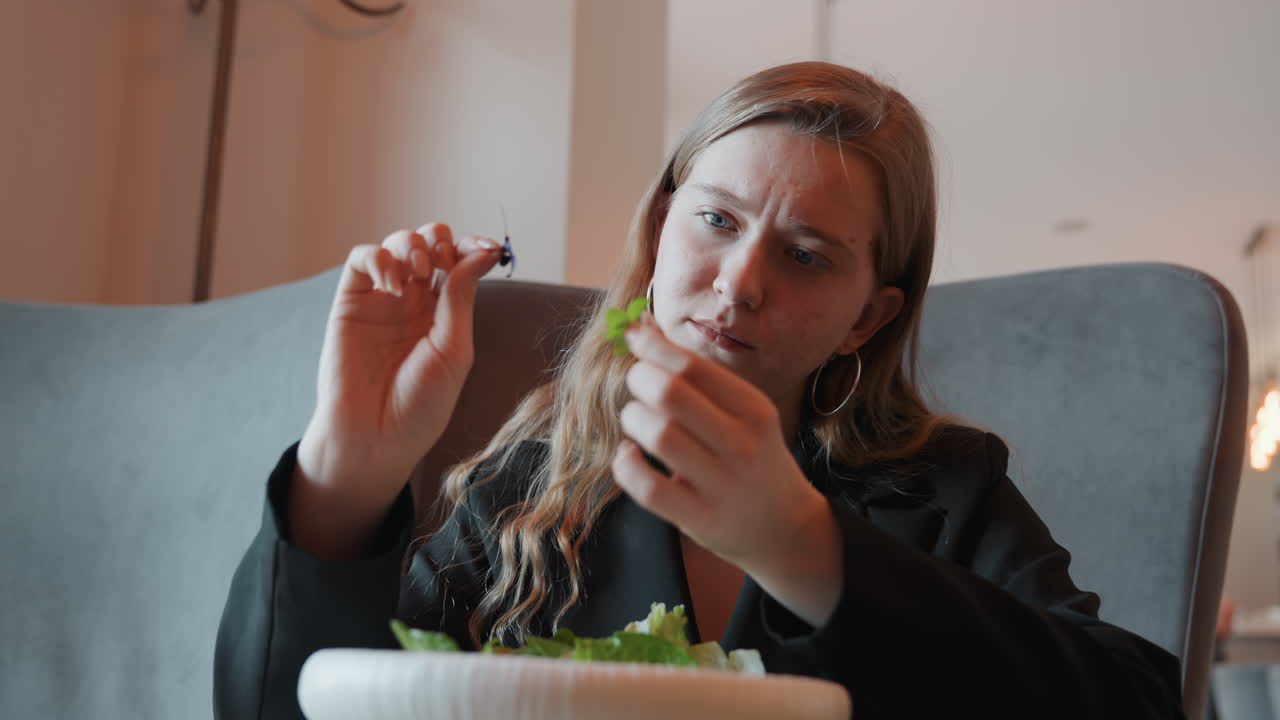 Weiße Frau prüft ein Salatblatt genau, Hände und Gabel im Bild, konzentrierter Ausdruck, Blazer und Creolen, Café-Interieur, wählerische Beurteilung von Textur und Frische, achtsame Essensszene.