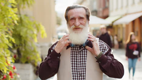 Happy-elderly-old-man-in-wireless-headphones-choosing,-listening-music-dancing-outdoors-city-street