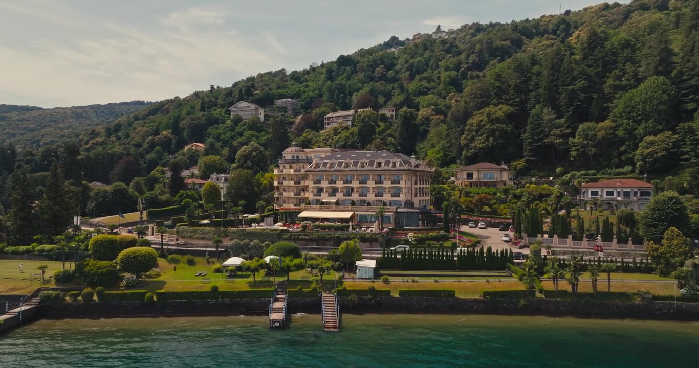 Drohne nähert sich einer historischen Villa am See, umgeben von üppigem Grün am Lago Maggiore. Türkisfarbenes Wasser, vorbeifahrende Autos und ein festgemachtes Boot schaffen eine malerische italienische Sommerszene