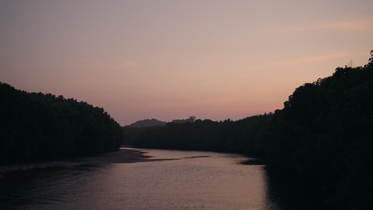 A serene Phuket river framed by dense mangrove forests glows in soft sunset hues, with calm water reflecting the fading pink-orange sky and distant hills