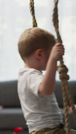 calm toddler boy rides on swing in light living room with sofa and bean bag chair. thoughtful blond boy holds on tight to ropes resting at home