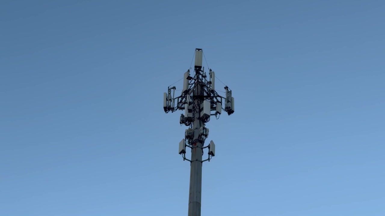 A tall telecommunications tower topped with multiple sector antennas, microwave dishes, and RF equipment, silhouetted against a soft blue evening sky.