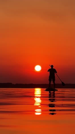 summer evening time on the calm river. lonely male silhouette on a sup board floating on water. orange sunset background. vertical video