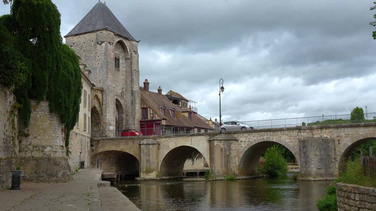 Histórico puente de piedra que cruza el río Loing en Moret-sur-Loing, enmarcado por la torre de una iglesia medieval, edificios a la orilla del río y un nublado paisaje rural francés