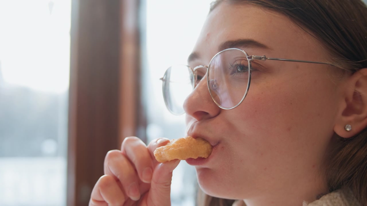 Young woman savoring fried snack with satisfied expression in sunlit cafe while seated by tall window, hand lifting bite near mouth, soft daylight shaping relaxed face and calm indoor mood