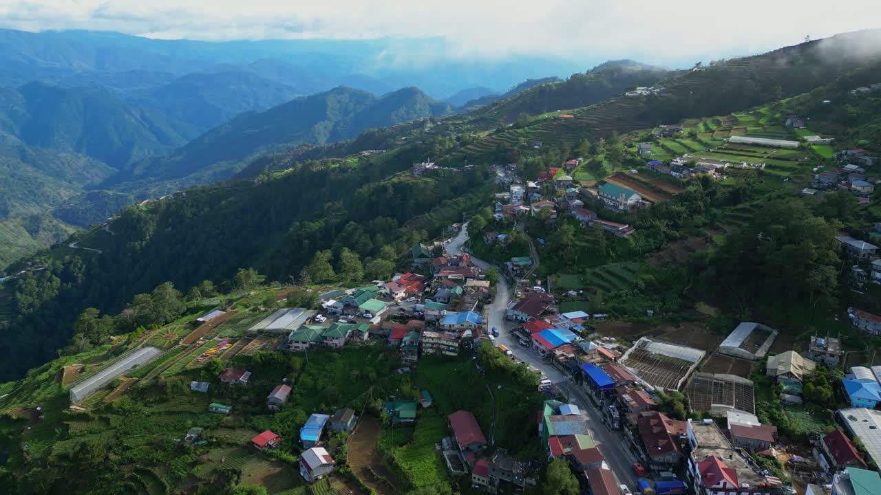 Plano aéreo abrangente e giratório sobre a rodovia sinuosa da montanha, mostrando a densa cidade à beira da estrada e os vibrantes terraços escalonados de Atok, Benguet, Filipinas