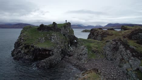 Man-standing-on-the-ancient-Dunscaith-Castle-ruins-historical-site-on-the-Isle-of-Skye-in-Scotland,-by-the-sea-on-a-cloudy-day