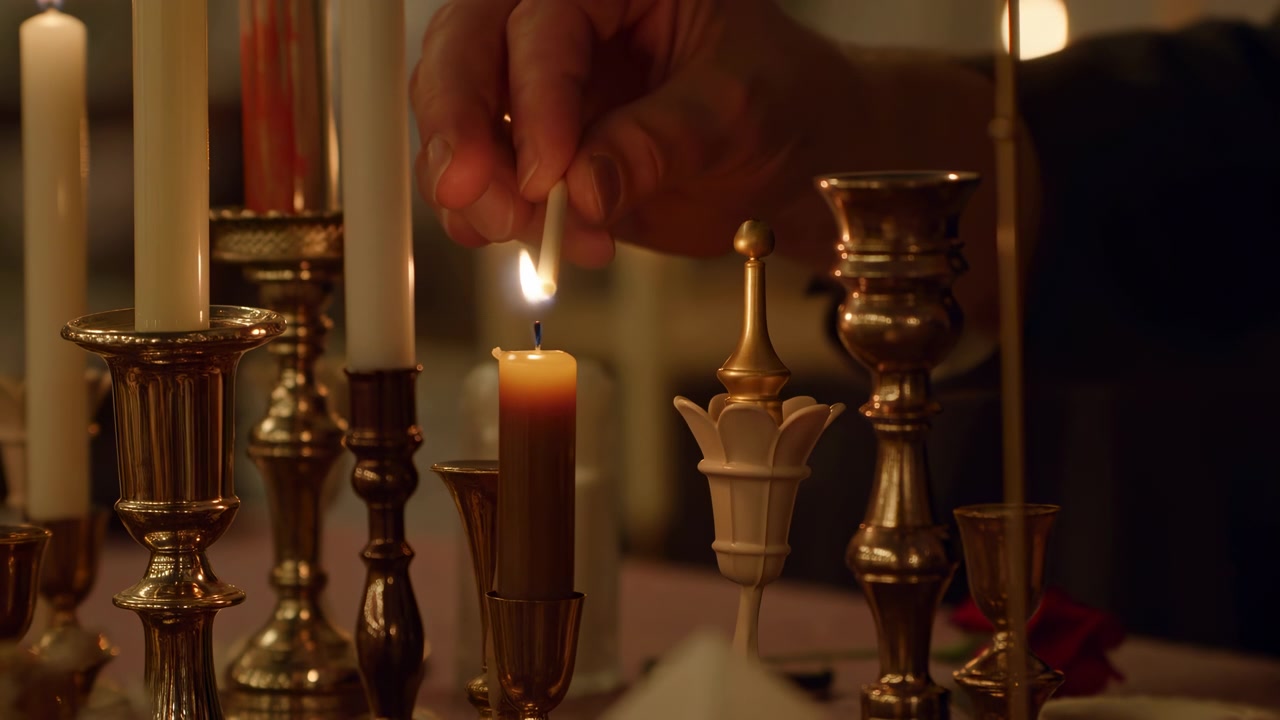 Nahaufnahme einer Hand, die ein brennendes Streichholz an den Docht einer braunen Stumpenkerze hält, wodurch eine gleichmäßige Flamme und ein sanfter Schimmer von geschmolzenem Wachs entsteht, inmitten von polierten Messingkerzenständern, hohen Stabkerzen, warmer Tischdekoration und edlem Ambiente.
