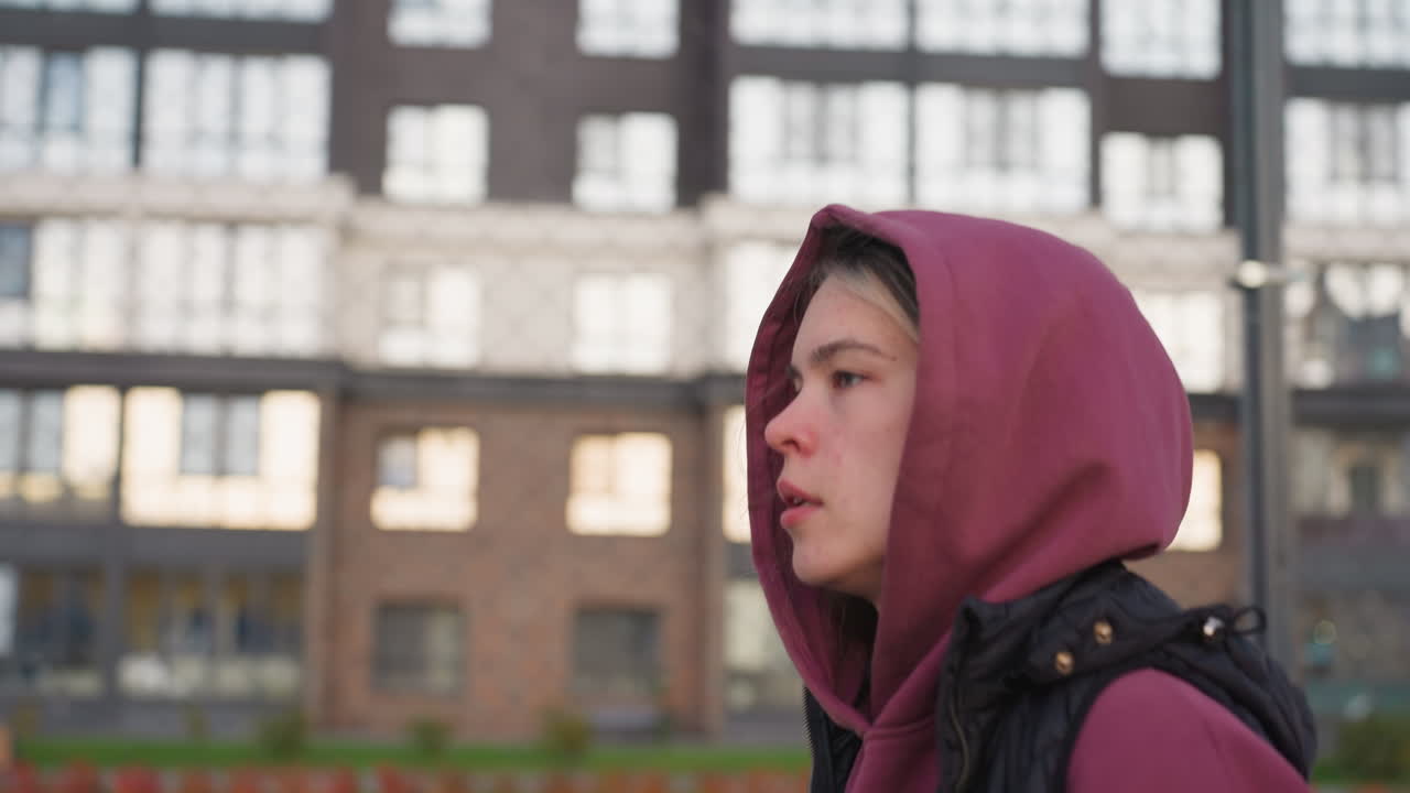Side view of strength trainer in hoodie jogging around curved outdoor sports court lined with barbed wire fence, silhouetted against soft dusk sky and urban apartment building backdrop