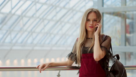 Hermosa-Chica-Hablando-Por-Un-Teléfono-Móvil-Mirando-A-La-Cámara-Y-Sonriendo.-Hermosa-Chica-Hablando-Por-Teléfono-Inteligente-Y-Sentada-Con-Bolsas-De-Compras-En-El-Centro-Comercial