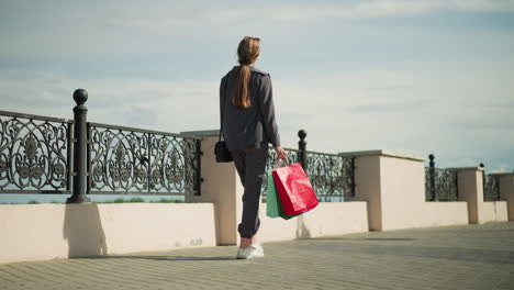 back view of lady in grey clothing carrying colorful shopping bags as she walks along an interlocked path near a decorative metal fence, outdoors on a sunny day, with shadows cast by the fence