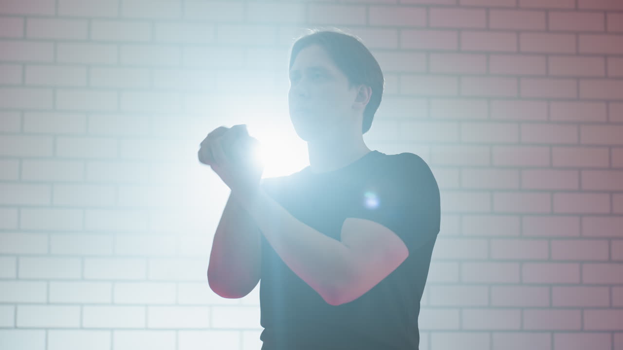Young man in black shirt warming up with serious focused expression against white brick wall background with bright backlight, symbolizing determination, preparation and readiness for action