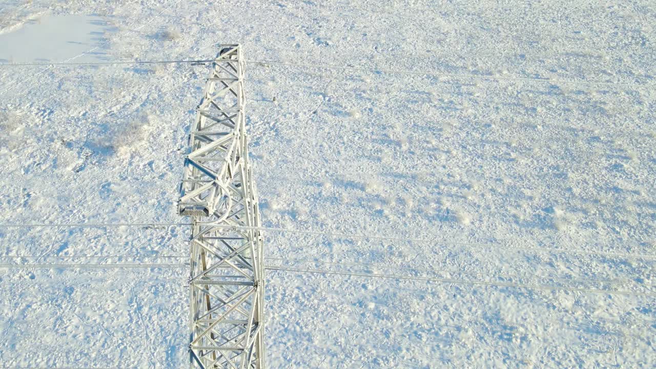 Ascending aerial movement around a frozen power line pylon reveals icy insulators and cables, ending in a top down view above a snow covered field under clear winter light