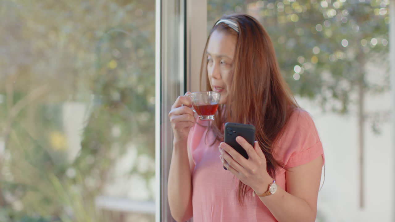 Holding smartphone and drinking tea, woman looking out window thoughtfully