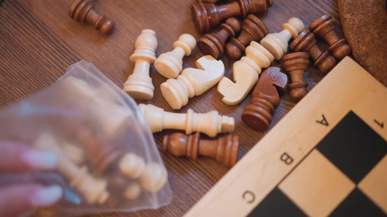 Close up of hand reaching into plastic bag to take chess pieces from wooden table with scattered white and brown pawns rooks visible. Wooden surface background includes partially seen